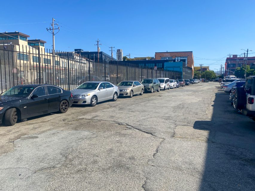 A row of parked cars lines one side of a street on a sunny day. Buildings and a tall fence are visible on the left side of the road.